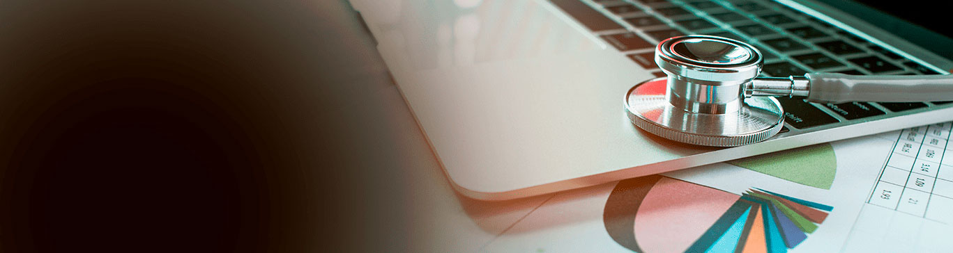 A ponta de um estetoscópio está sobre o teclado de notebook, por cima de relatórios com gráficos coloridos.