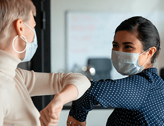 Duas pessoas do sexo feminino em cumprimento mútuo com o cotovelo. A iniciativa atende pacientes que receberam alta hospitalar e persistem com manifestações clínicas da doença.