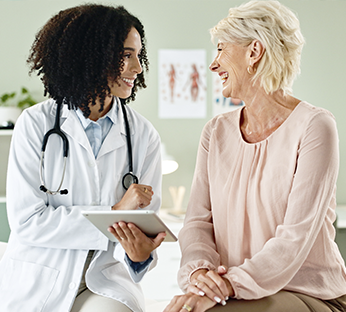 Médica sorridente conversando com uma paciente idosa em um consultório, enquanto segura um tablet.