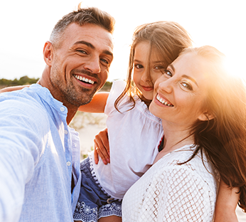 Família sorridente, composta por um homem, uma mulher e uma menina, tirando uma selfie ao ar livre em um dia ensolarado.