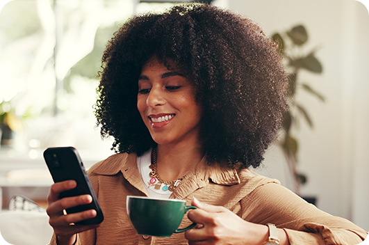 Mulher sorrindo enquanto usa o celular e segura uma xícara de café.
