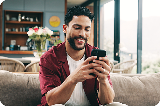 Homem sorrindo enquanto usa o celular sentado no sofá de casa.