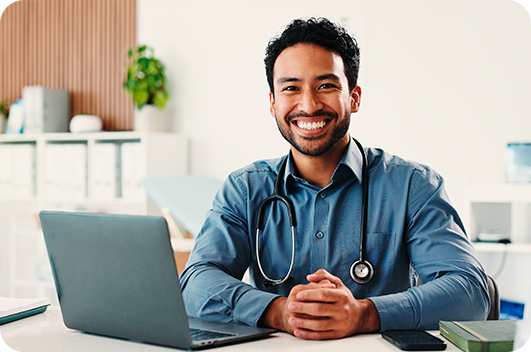 Médico sorrindo em consultório com notebook e estetoscópio no pescoço.