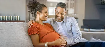 Casal sentado juntos no sofá, felizes e contemplando barriga em avançada gestação.