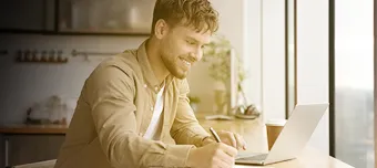 Homem sorridente em frente à janela, com notebook aberto e escrevendo em caderno.