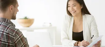 Moço sentado em frente a uma mesa, sendo atendido por moça profissional sorridente apresentando documento.
