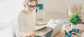 Pessoa trabalhando na escrivaninha em frente ao computador e segurando documentos.