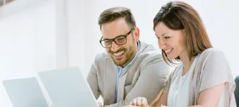 Casal sorridente, sentados à mesa e apontado telas dos notebooks.